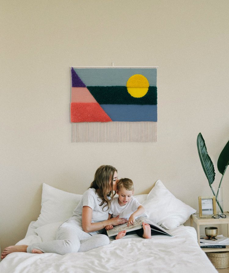 Opera tessile contemporanea tessuta a mano da Ilary Bottini, paesaggio astratto con toni di azzurro, blu, verde, rosa, viola e giallo con campiture geometriche orizzontali, diagonali e circolari a alternanza di sezioni tessute e annodate, appeso in una stanza bianca con una donna e un bambino che leggono su un letto