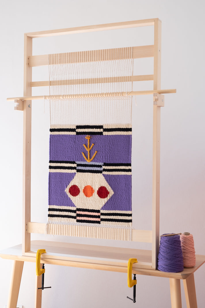 Telaio verticale in legno massello per tessitura di tappeti e arazzi, montato su un tavolo in legno, ordito e con opera incompiuta con base viola bianca e nera e elementi rossi, arancioni e senape