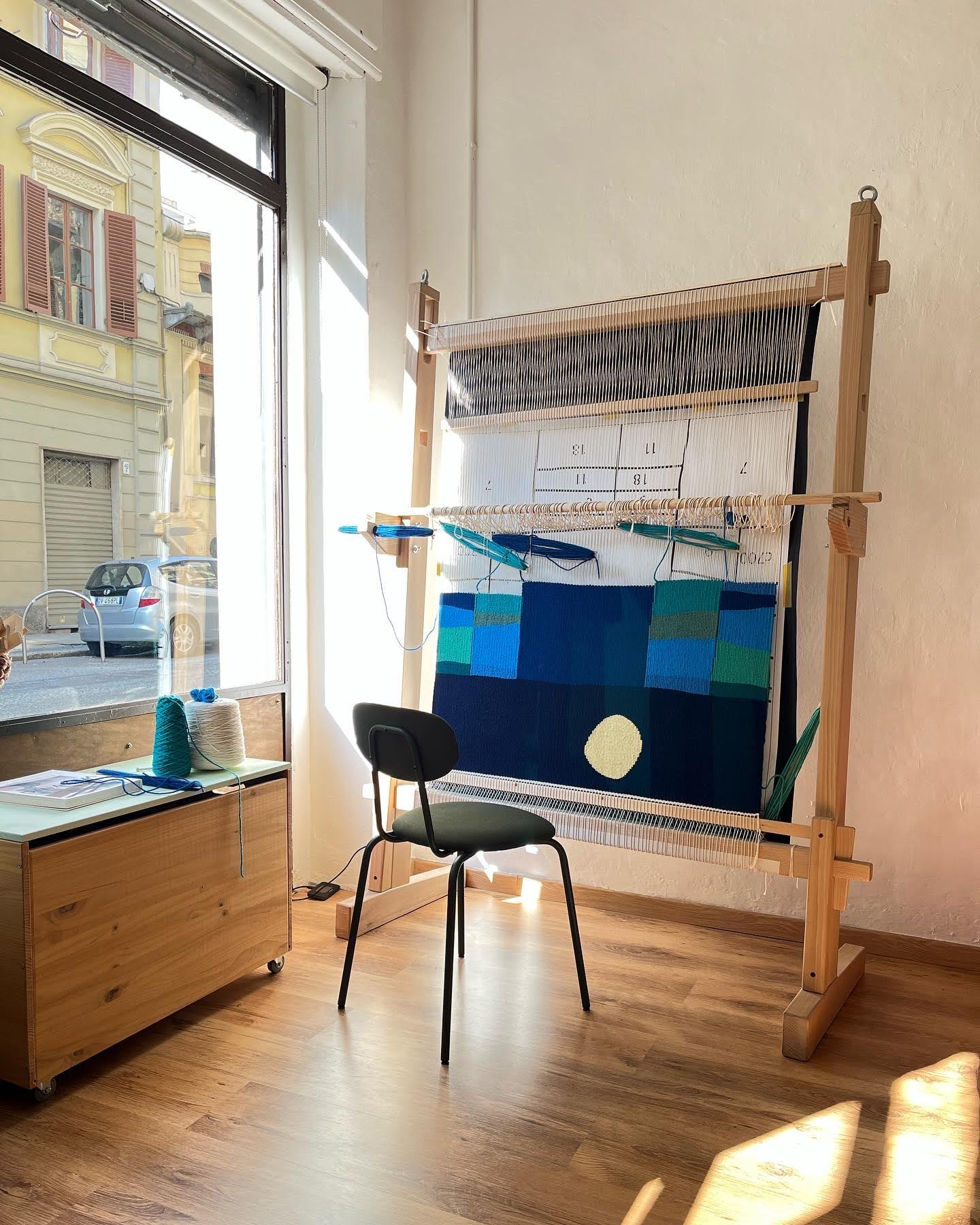 Studio dell'artista tessile Ilary Bottini, telaio da tessitura verticale con opera work in progress raffigurante un paesaggio marino astratto notturno con toni di blu, verde e giallo, di fianco a una vetrina su strada