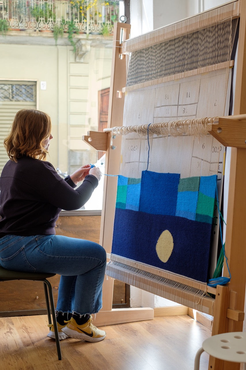 Artista tessile Ilary Bottini seduta davanti a un telaio verticale mentre tesse un'opera astratta in lana raffigurante un paesaggio marino notturno con toni di blu, verde e giallo 