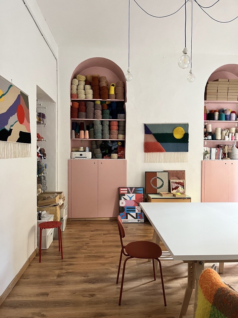 Studio di fiber art di Ilary Bottini, stanza bianca con nicchie, mensole e armadi rosa, rocche di lana sarda in vari colori, opere di fiber art e arazzi contemporanei appesi alle pareti