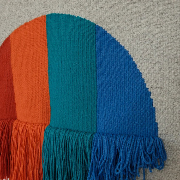 Arazzo contemporaneo in lana su base in cotone di Ilary Bottini con forma semicircolare e campiture verticali nei toni rosso, arancio, verde e blu, con frange decorative nella parte inferiore.